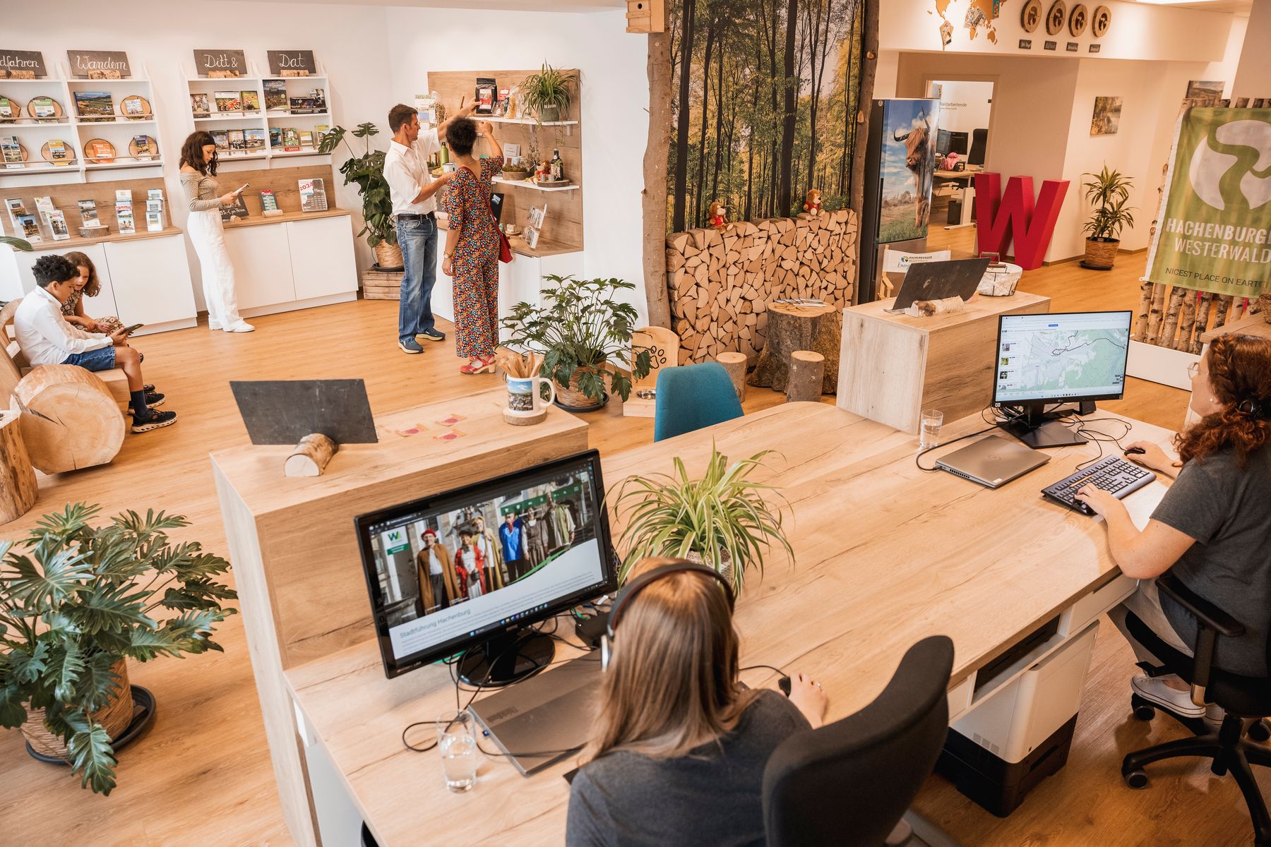 Modernes Büro mit Holzdekor und Pflanzen. Im Vordergrund arbeiten zwei Personen an Computern, während mehrere andere in der Nähe von Regalen mit Broschüren und Produkten im Hintergrund interagieren. Der Raum wirkt offen und einladend.